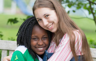 Two girls sit and smile at the camera. Promotional photo for summer 2017 Home's Grown Kids Newsletter.